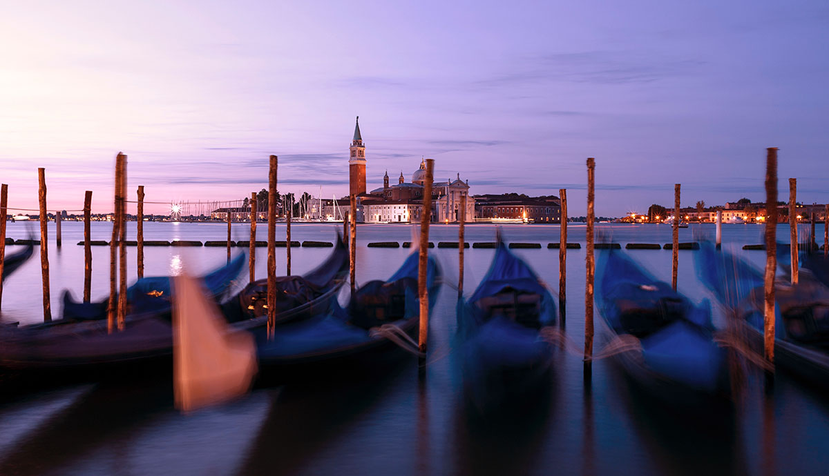 Blurry image of gondolas docked on a body of water in Italy in the evening | Image Credit: Geoffroy Hauwen via Unsplash