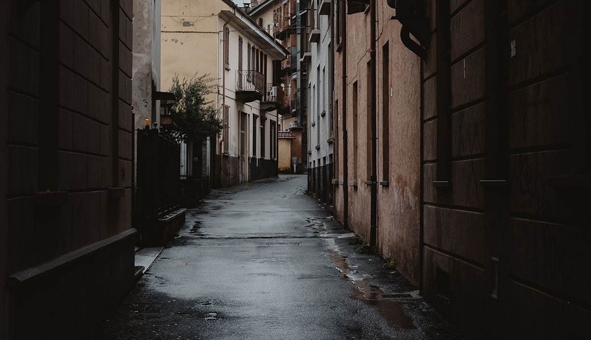 Header Graphic: dark and rainy street tunneled by tan buildings and copper brick buildings that curve right into the crowded distance| Image Credit: Unsplash