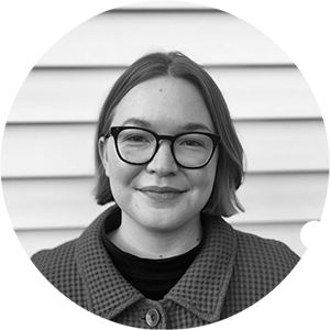 Author Abby Waters stands in front of the siding of a building in a black and white picture, wearing glasses and a waffle knit shirt with hair falling just below her chin