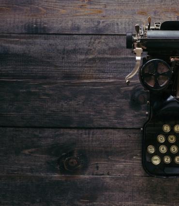 Header Graphic: Typewriter against a dark, wooden desk | Image Credit: Patrick Fore